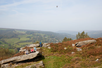 Balloon over Curbar Edge This landscape photograph, taken in the late morning during autumn, shows a hot air balloon floating over Curbar Edge in the Peak District, England. The image captures the rural scenery of the United Kingdom, featuring the striking rocky outcrops of Curbar Edge overlooking expansive green valleys and woodlands. In the foreground, several people are seated on the large stones, enjoying the scenic views typical of the Peak District. The rolling hills and patchwork fields exemplify the rural charm of this area of England, making Curbar Edge a well-known viewpoint in the region.